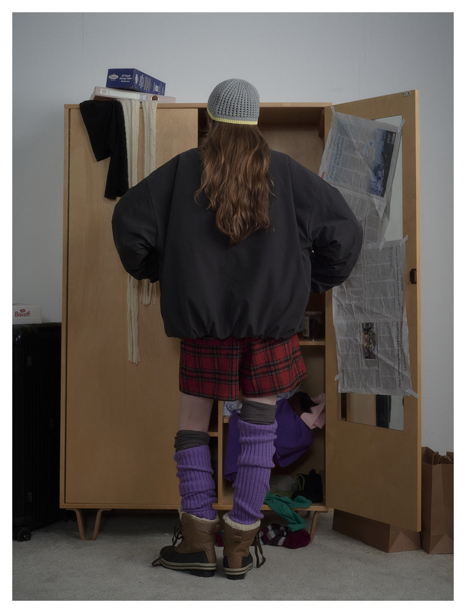 Rear view of a model in an oversized black jacket, red plaid shorts, a gray knit hat, and purple leg warmers.