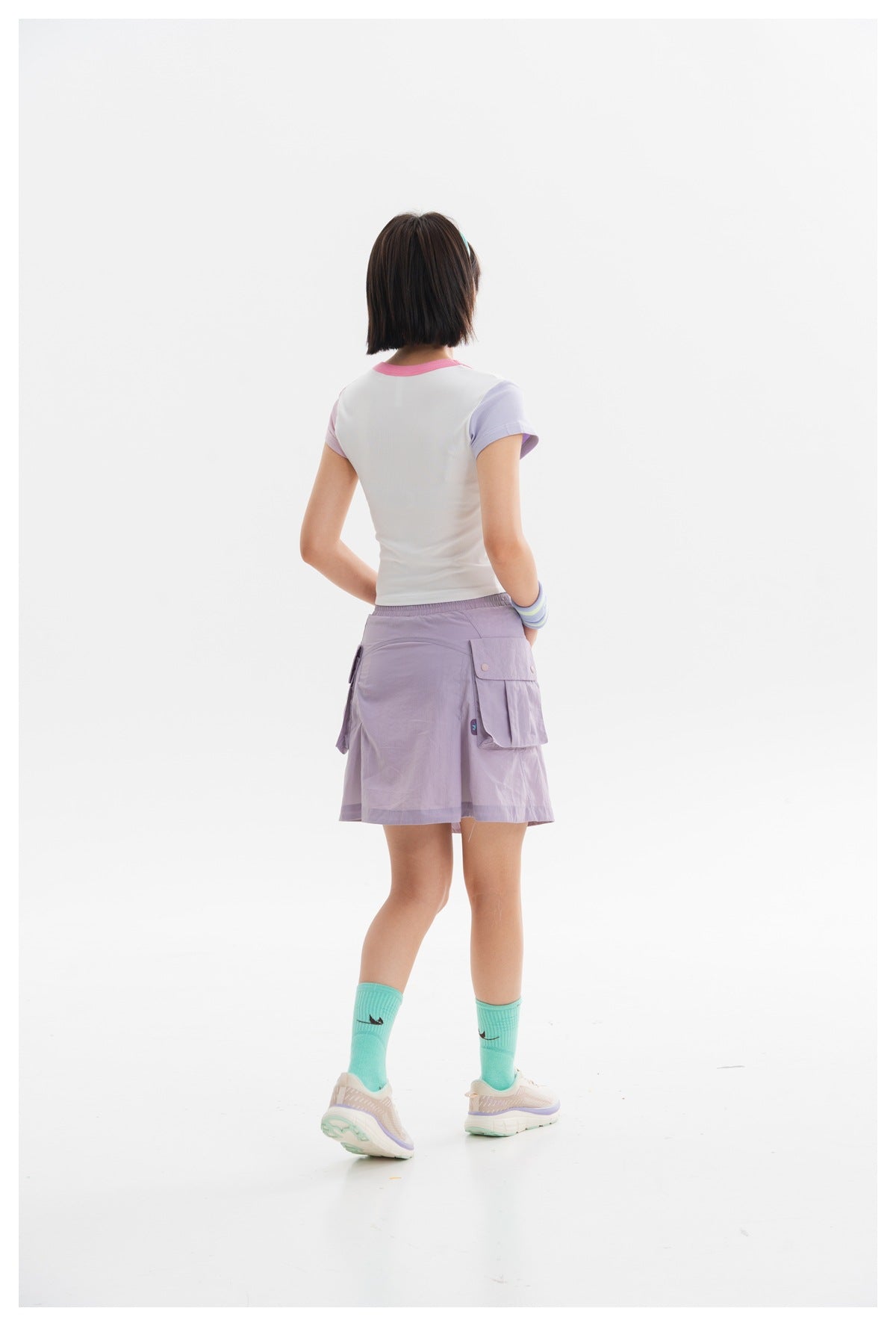 Rear view of a model wearing a white crop top with pink neckline and lilac sleeves, and a lavender cargo skirt.