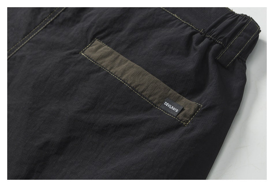 Close-up of dark pants with an olive-gray pocket strip, gathered side waistband, contrast stitching, and a small sewn label.