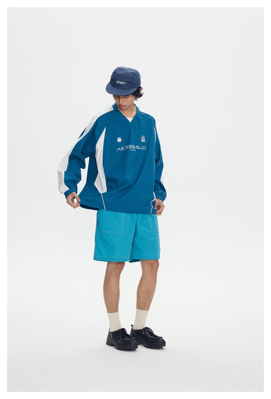 Model wearing a blue-green V-neck pullover with white sleeve panels and teal shorts, shown from the front.