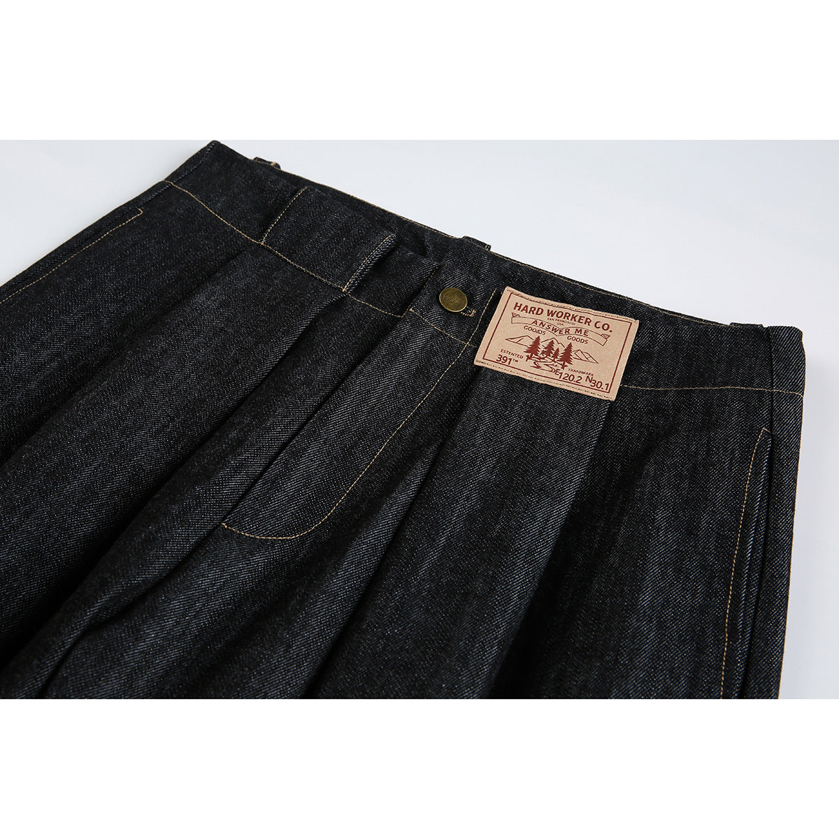 Close-up of dark indigo jeans showing the waistband, pleats, brass button, tan contrast stitching and sewn leather-like