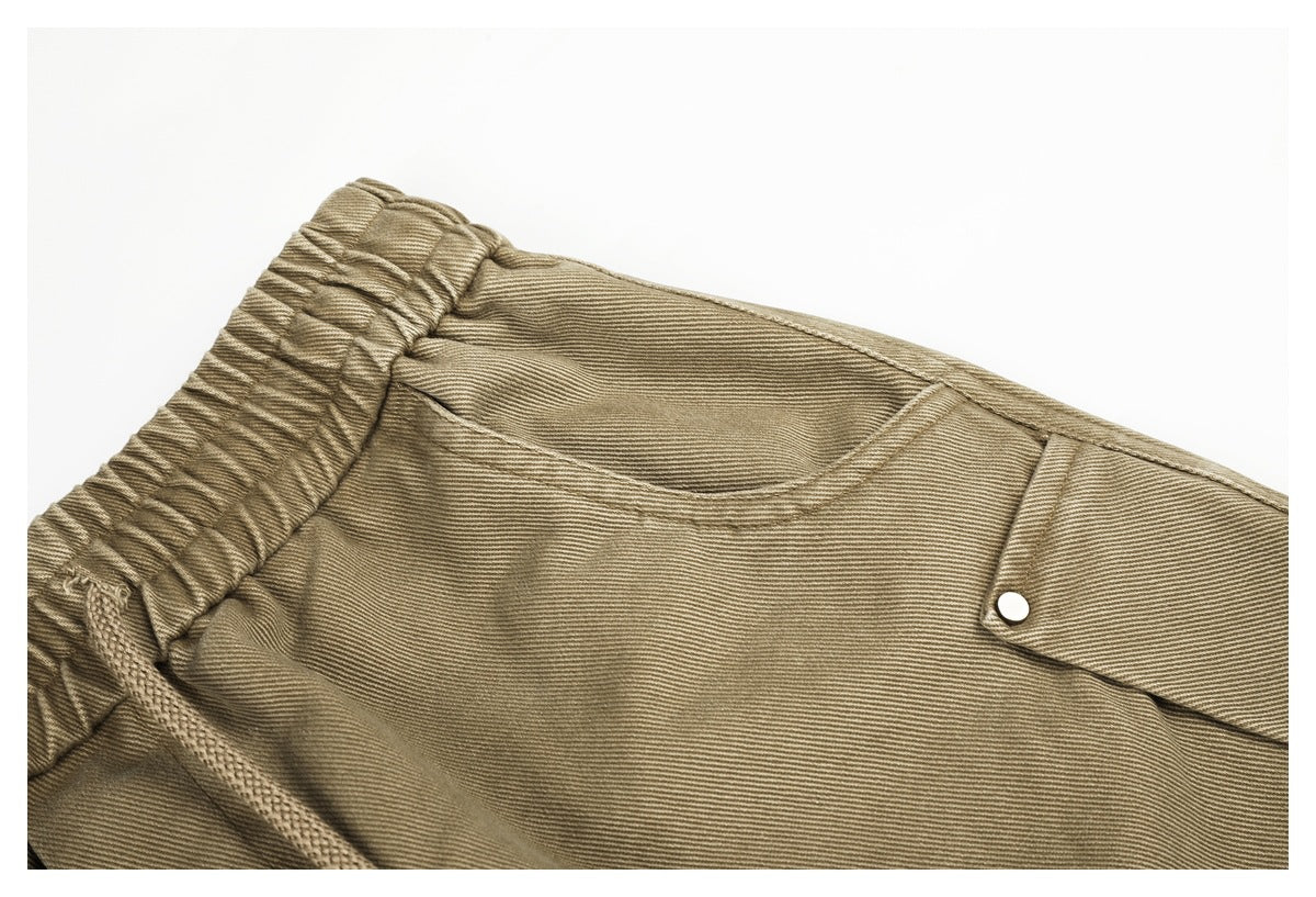 Close-up of khaki trousers showing the gathered elastic waistband, curved pocket, drawstring, and snap-button flap pocket.