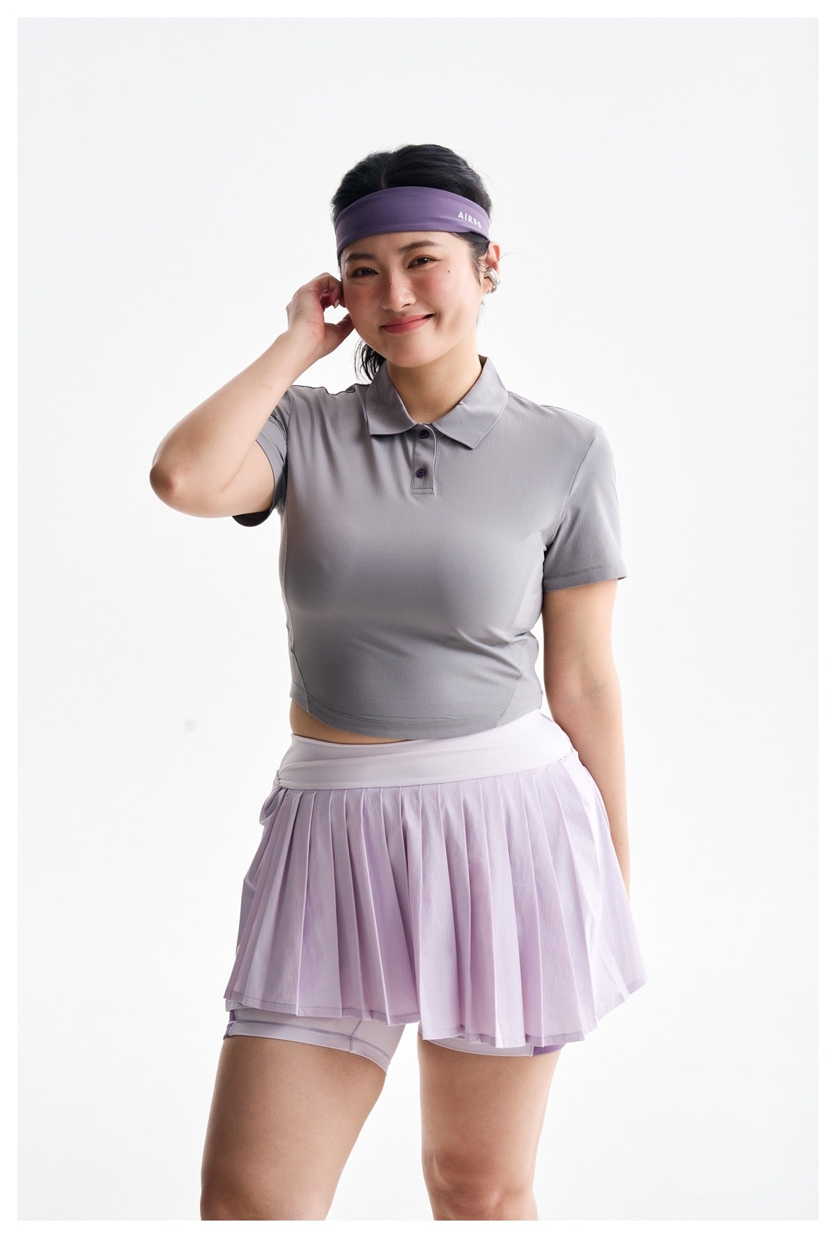 A woman models a gray cropped polo and lilac pleated skort, adjusting an earbud; her purple headband has small white
