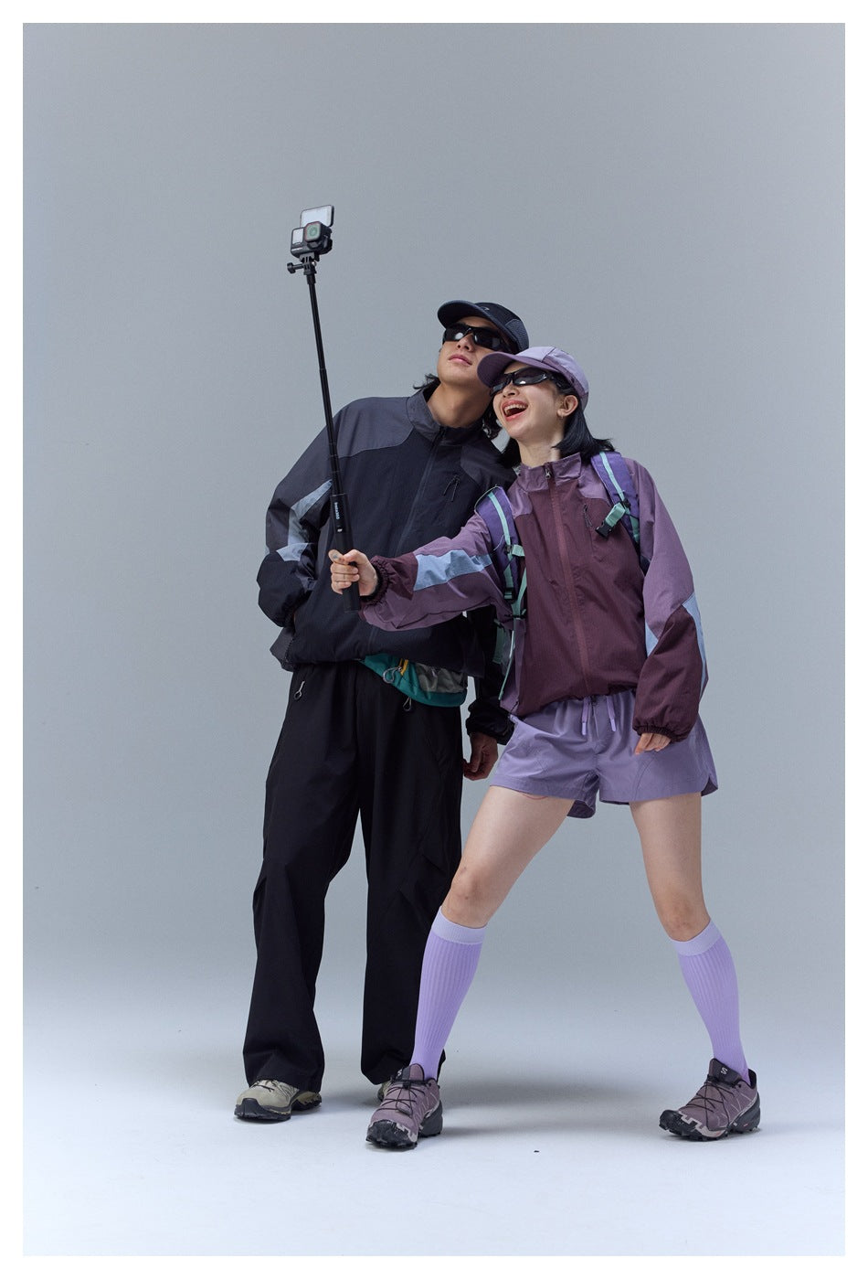 Man and woman wearing contrasting color-block jackets; the woman holds a selfie stick in a studio setting.