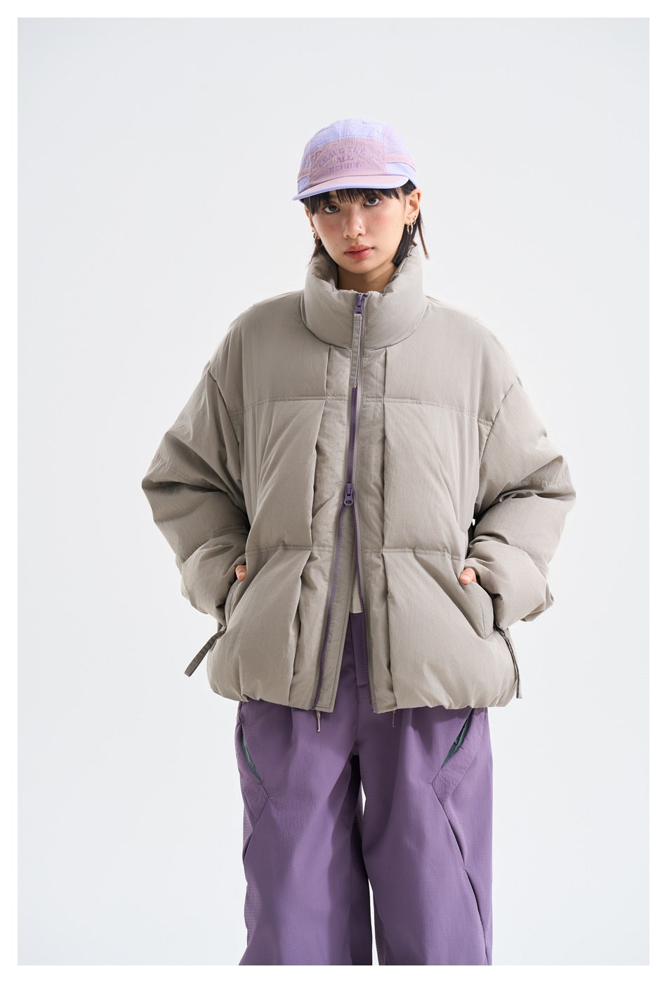 Closer front view of a model in a light gray puffer jacket with a high collar and purple trousers.