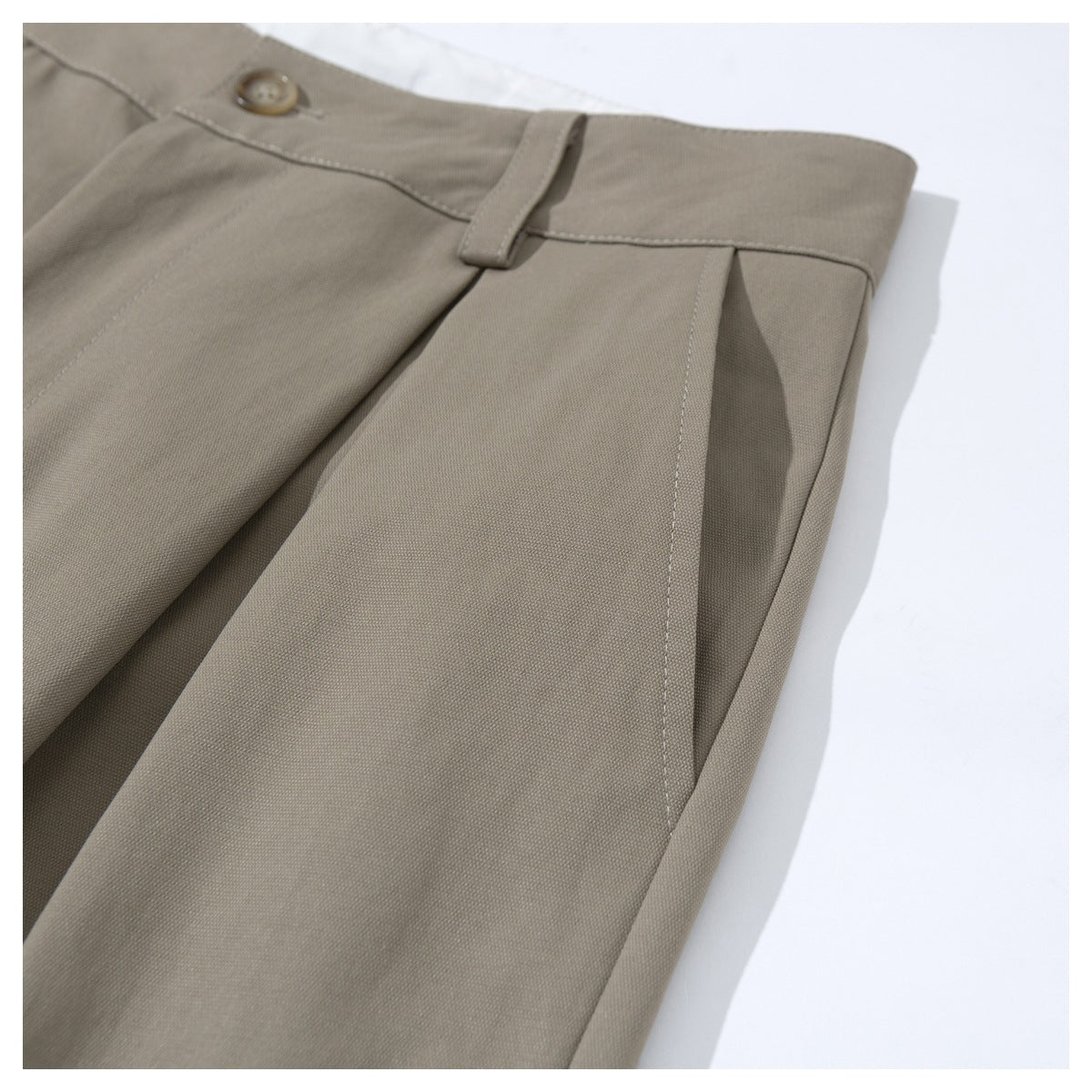 Close-up of taupe trousers showing the waistband, button, belt loop, and front pleat.