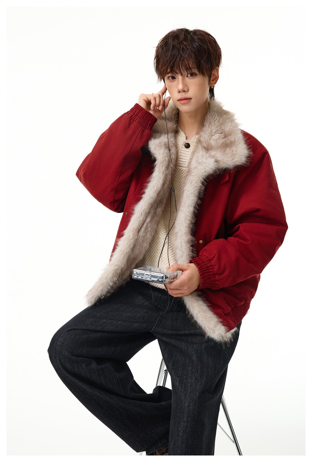 Seated model wearing an open deep red jacket with a wide beige fur-trimmed collar and dark jeans.