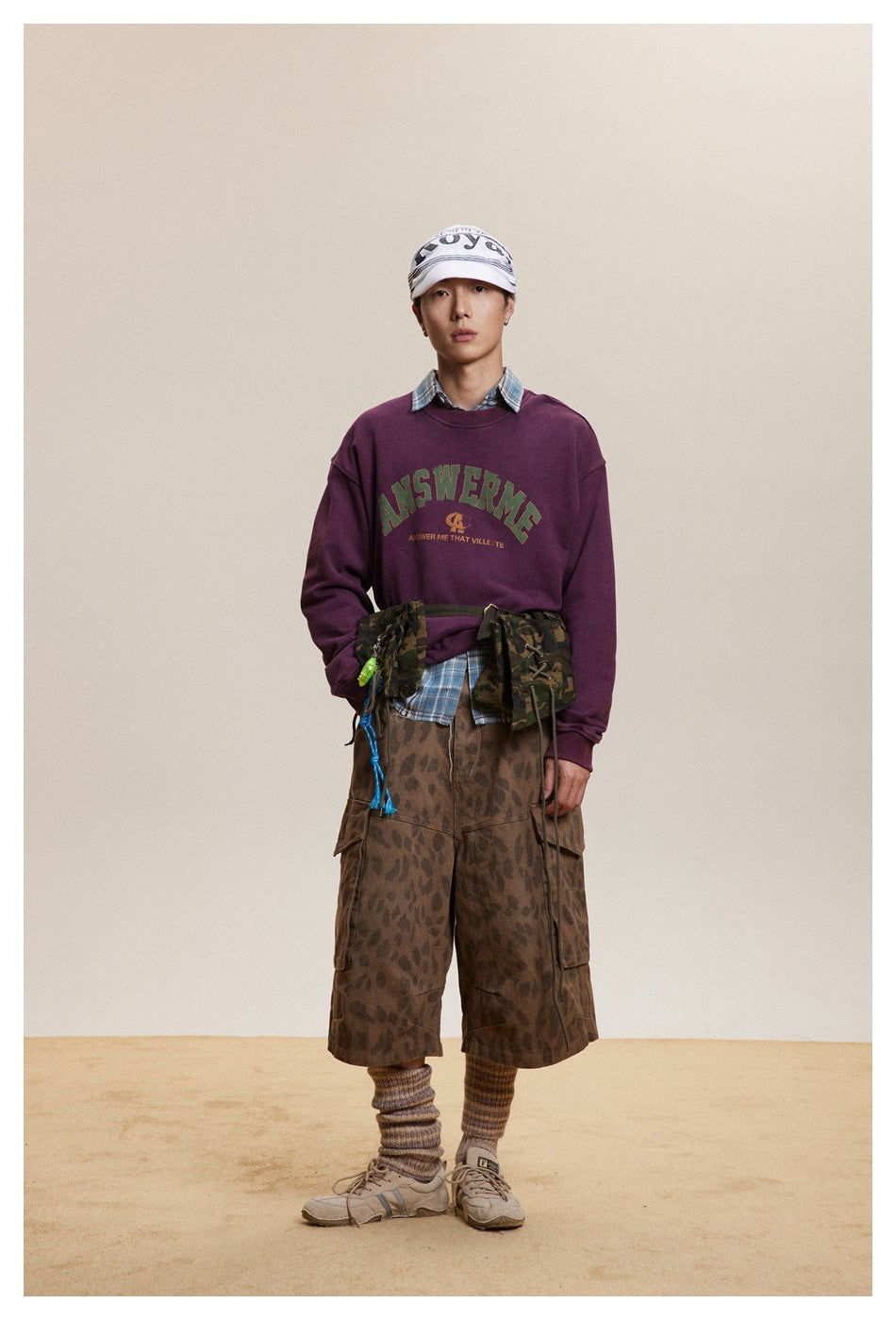 Model wearing a burgundy graphic crewneck sweatshirt over a plaid shirt and brown patterned cargo shorts.
