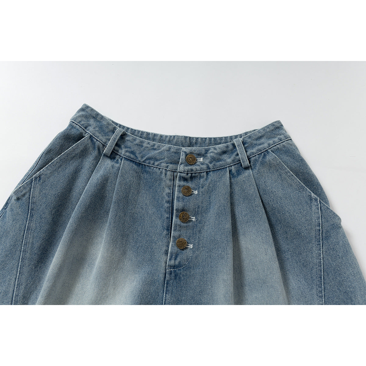 Close-up of blue denim trousers showing the waistband, pleats, belt loops and four brass-tone buttons.
