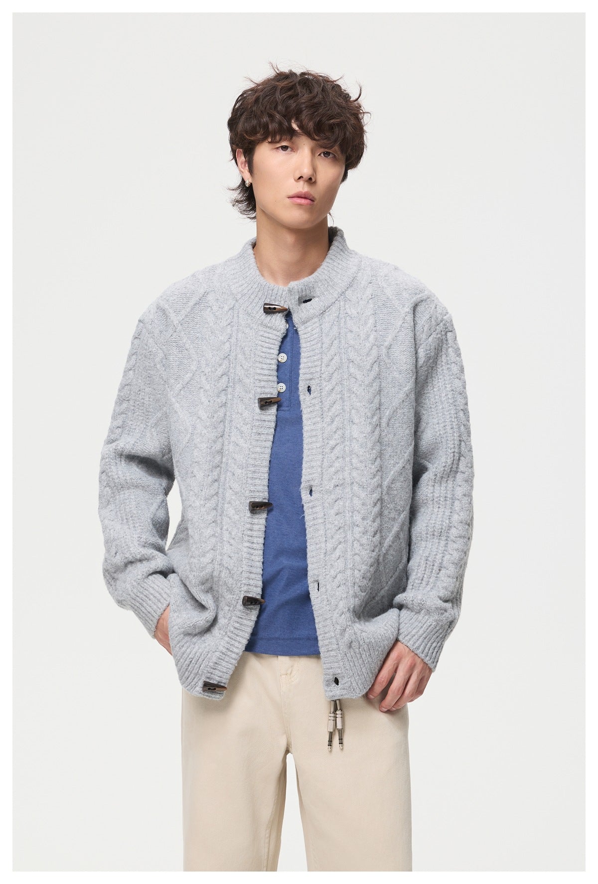 Model wearing an open light gray cable-knit toggle cardigan over a blue shirt and cream trousers.
