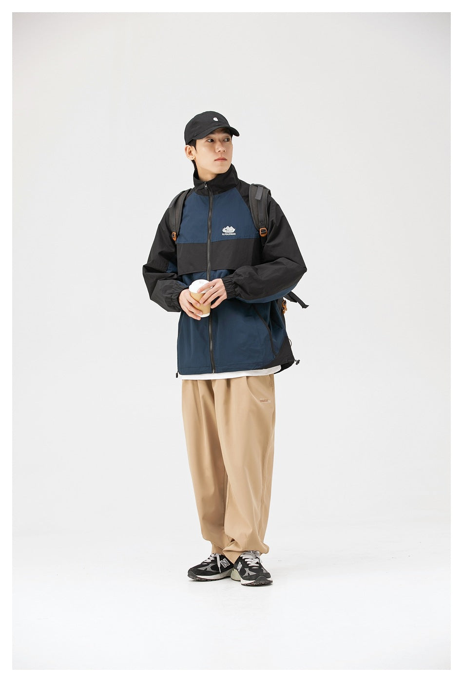Full-length three-quarter view of a model wearing a navy and black color-block zip jacket and beige trousers.