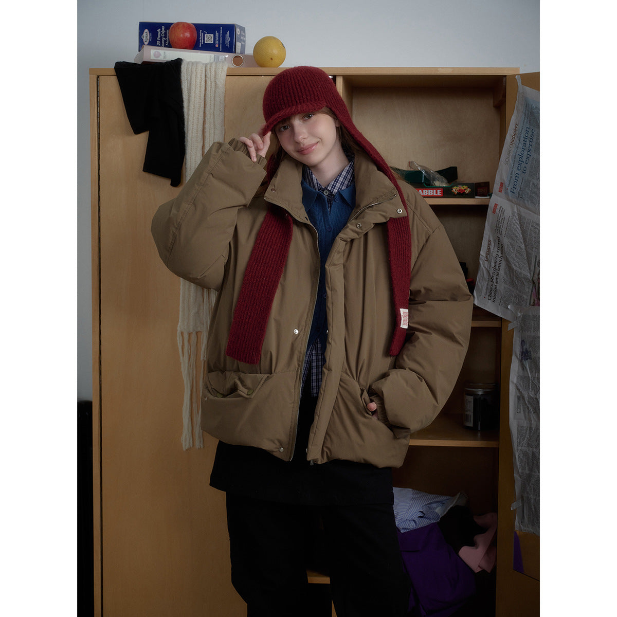 Closer view of a model in a brown puffer jacket and burgundy knit hat and scarf, layered over a blue vest and checked shirt.