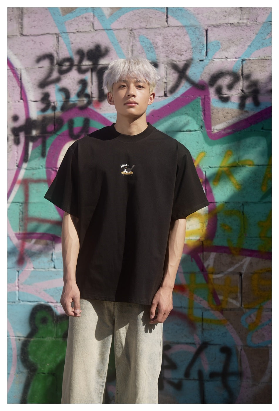 Front view of a model wearing a black crew-neck T-shirt with a small illustrated chest graphic against a colorful graffiti