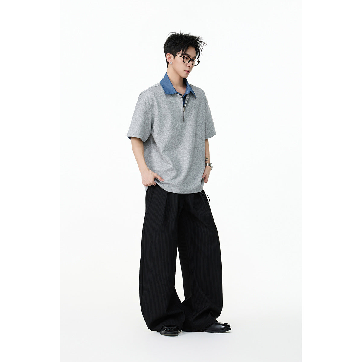 Model wearing a loose gray short-sleeve quarter-zip polo with a blue collar and wide black trousers.