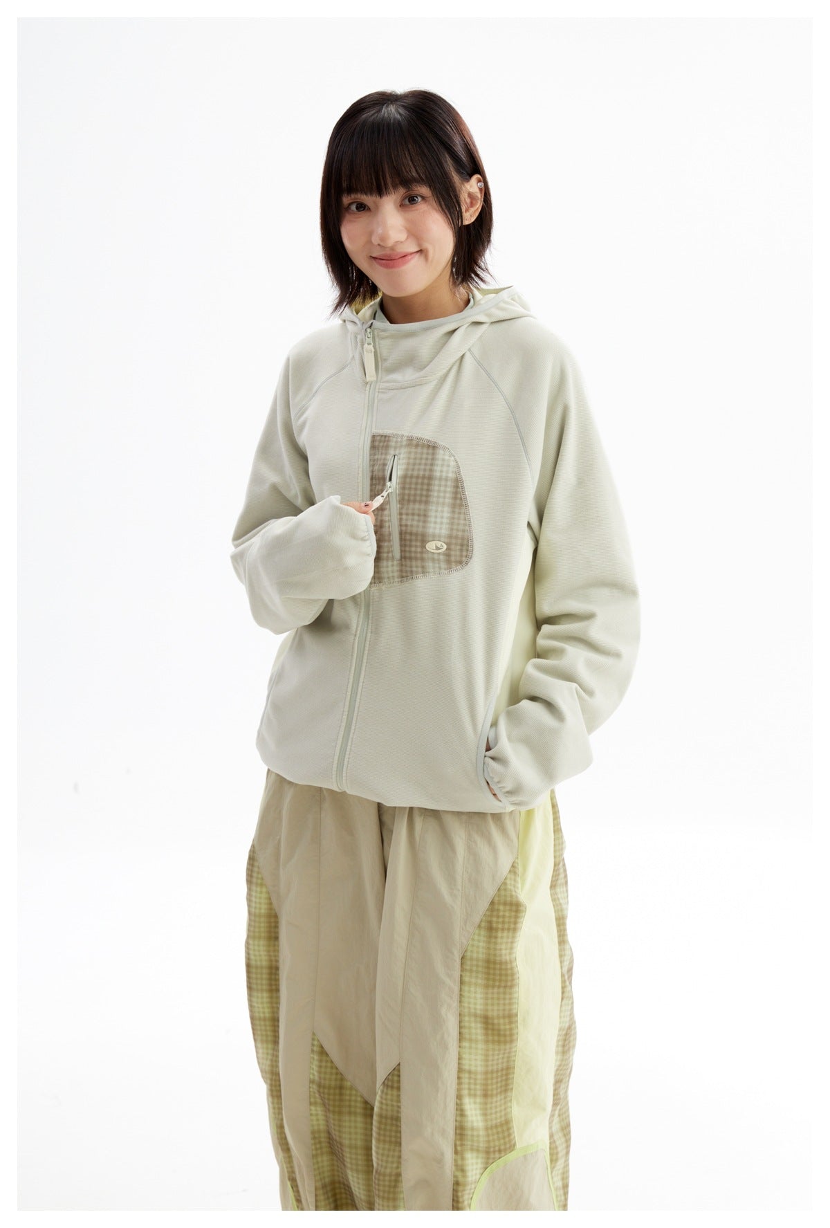 Model wearing an ivory hooded zip jacket with a checked chest pocket and loose beige-and-green checked trousers.