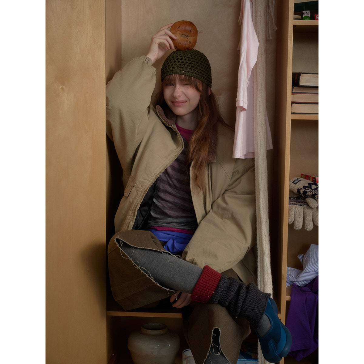 Model seated in a closet wearing an oversized beige jacket with a brown collar and a dark olive crocheted beanie.