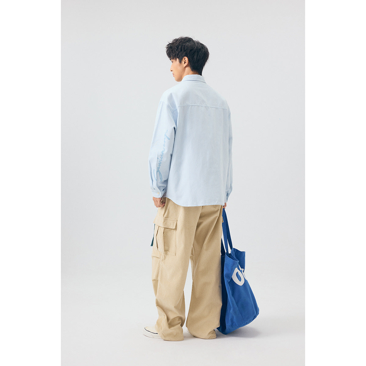 Rear view of a model in a pale blue shirt with sleeve lettering and beige cargo pants, carrying a blue tote.