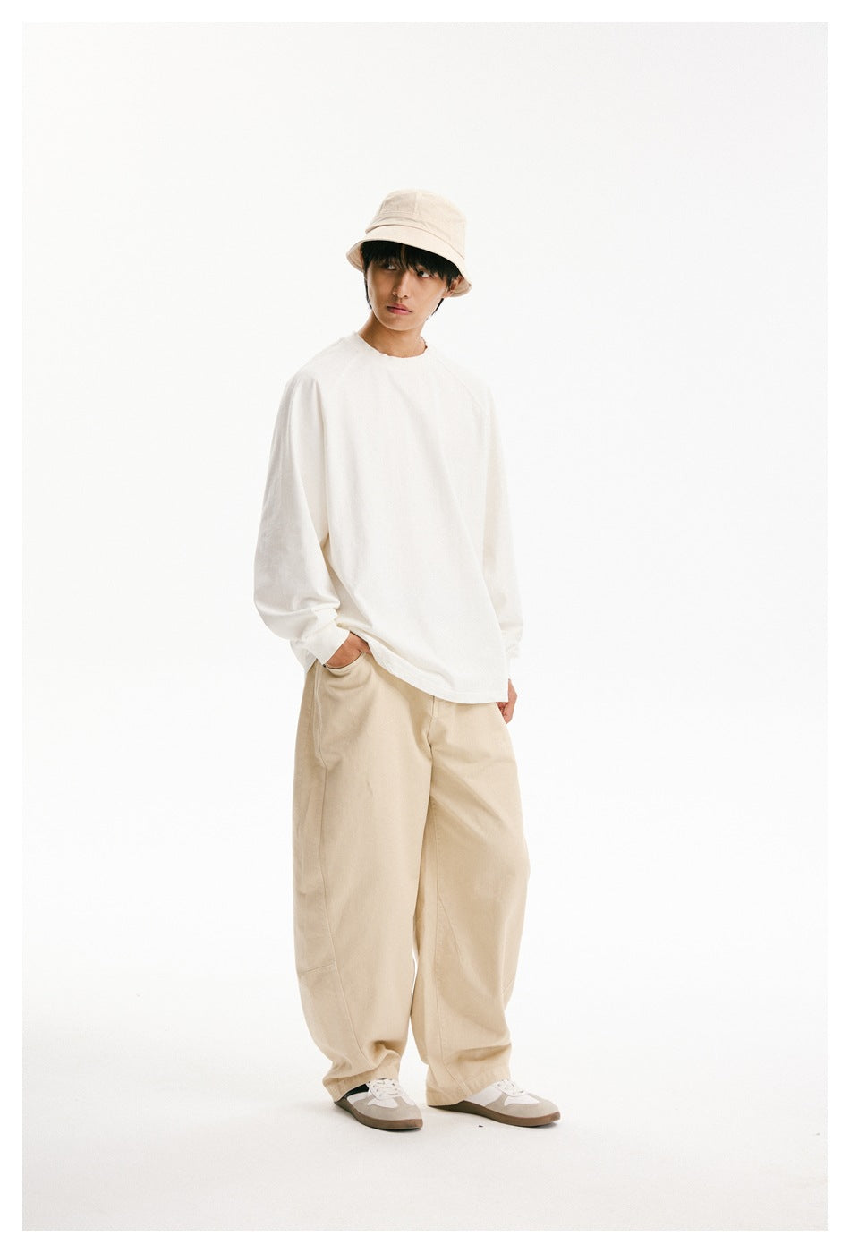 Angled full-length view of a model wearing a loose white long-sleeve shirt and beige trousers with a bucket hat.