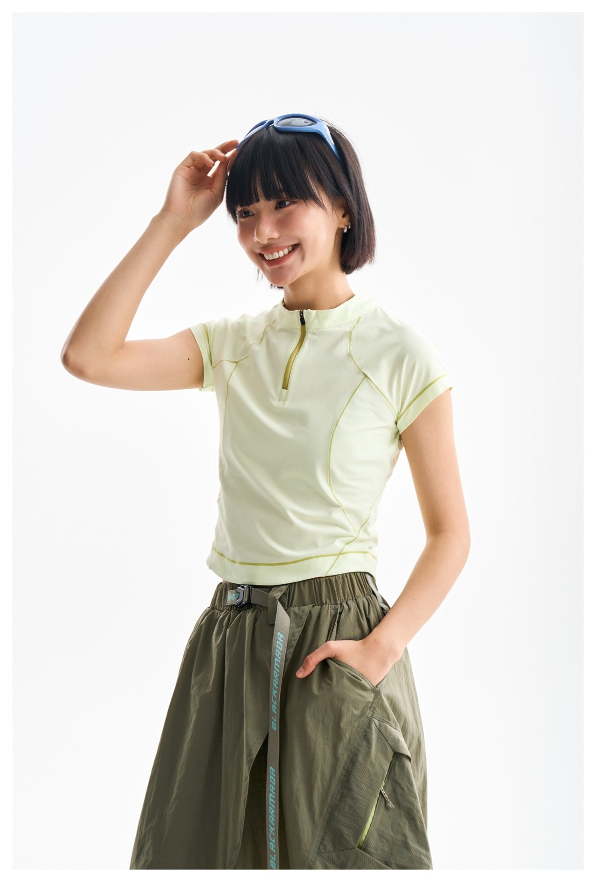 Model in a pale yellow zip-neck top and olive skirt, adjusting her blue sunglasses in a three-quarter view.