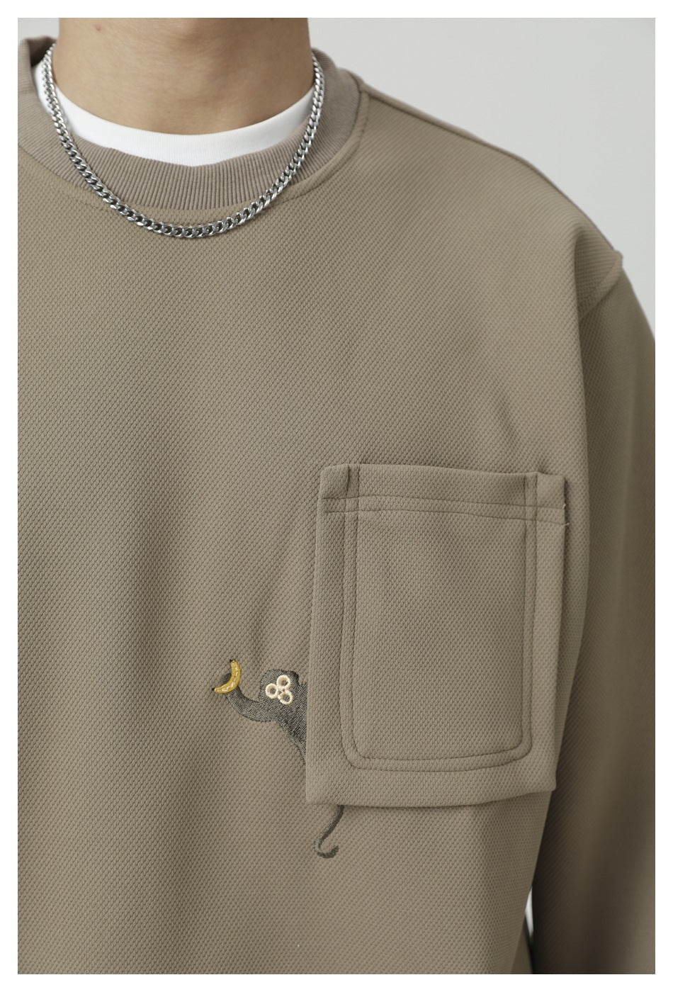 Close-up of the taupe sweatshirt’s textured fabric, ribbed neckline, chest pocket, and small embroidered motif.