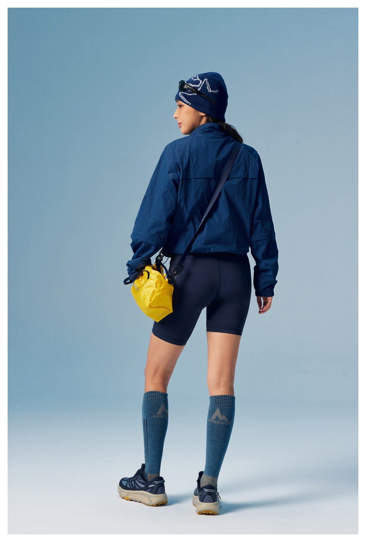 Rear view of a model wearing a navy jacket and dark shorts with blue knee socks; a yellow bag hangs at her side.