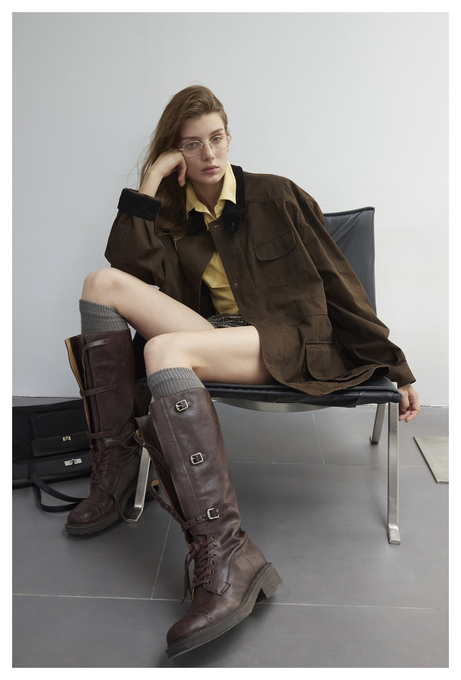 Seated model wearing a dark brown oversized jacket, yellow shirt, plaid skirt, and tall brown buckle boots.