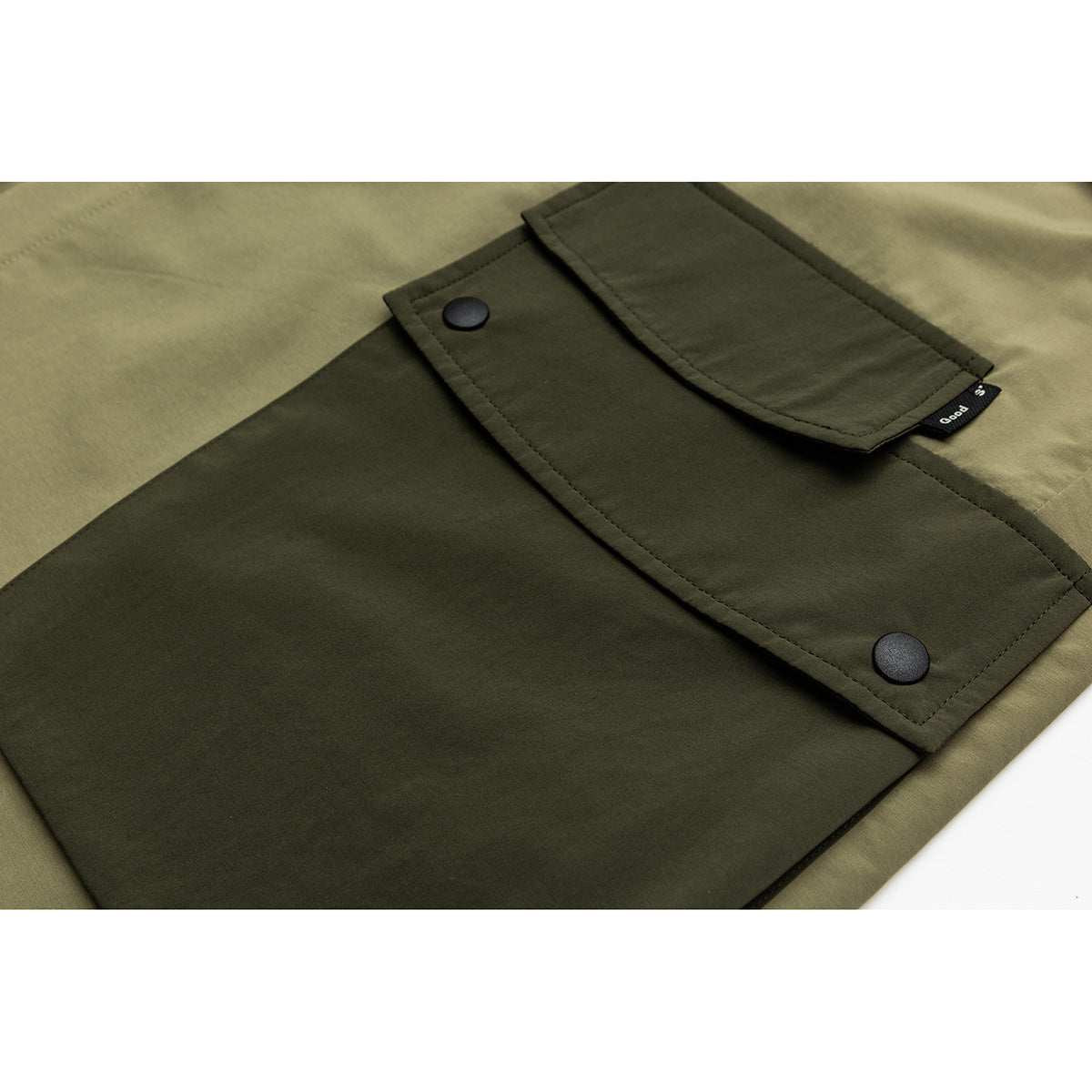 Close-up of an olive garment's dark patch pocket with a flap, two black snaps, and a small side tag.
