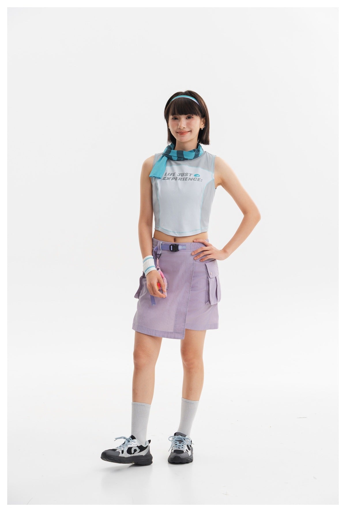 Model wearing a pale blue sleeveless crop top and lavender wrap-front cargo skirt with a striped scarf.