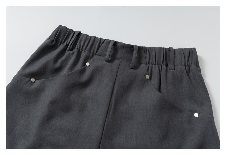 Close-up of charcoal trousers showing an elastic waistband, belt loops, curved pockets, and small metal studs.