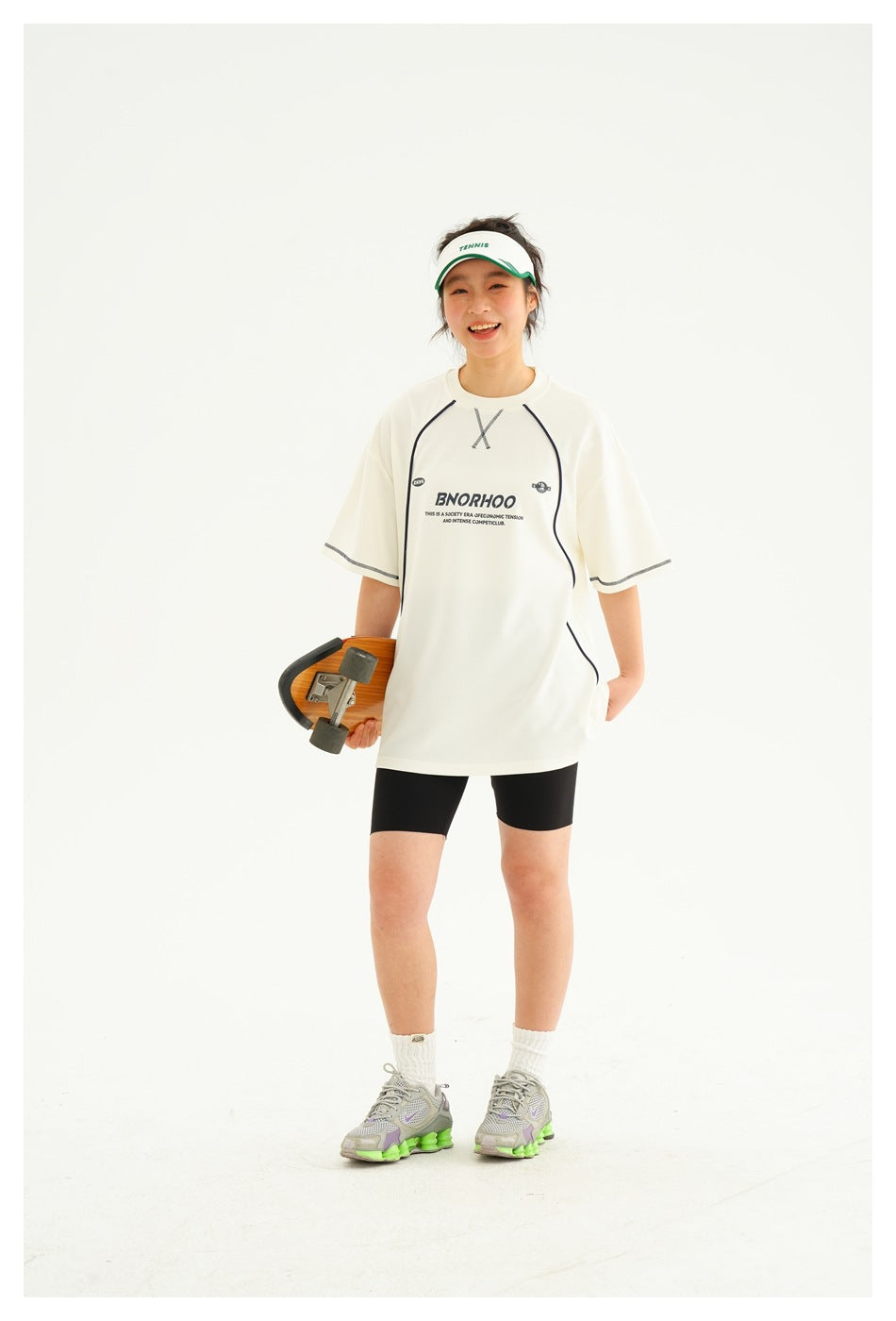Full-length model wearing an ivory oversized graphic T-shirt with dark piping and black shorts, holding a skateboard.