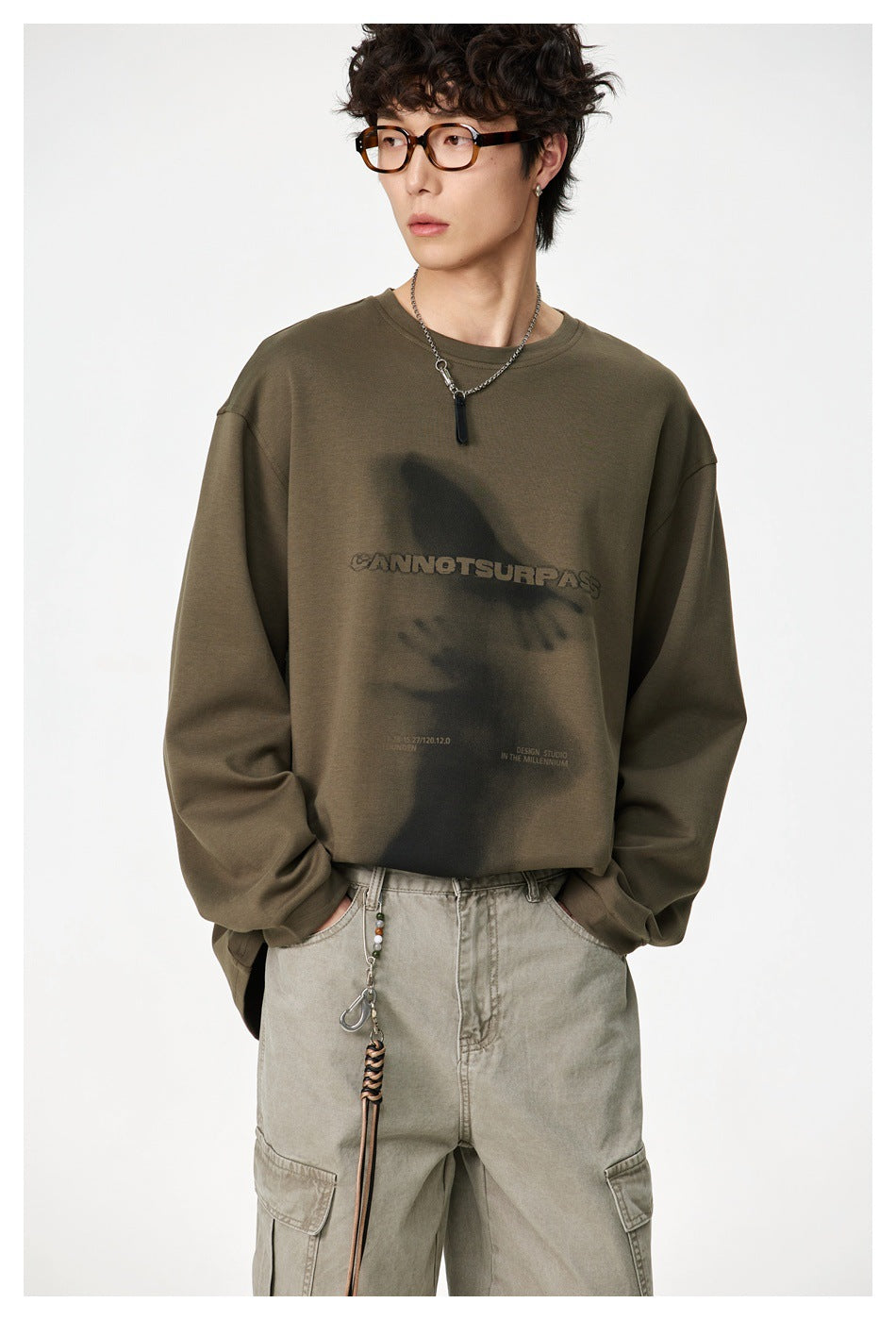 Model wearing an olive oversized sweatshirt with a dark graphic and text print, paired with light cargo pants.