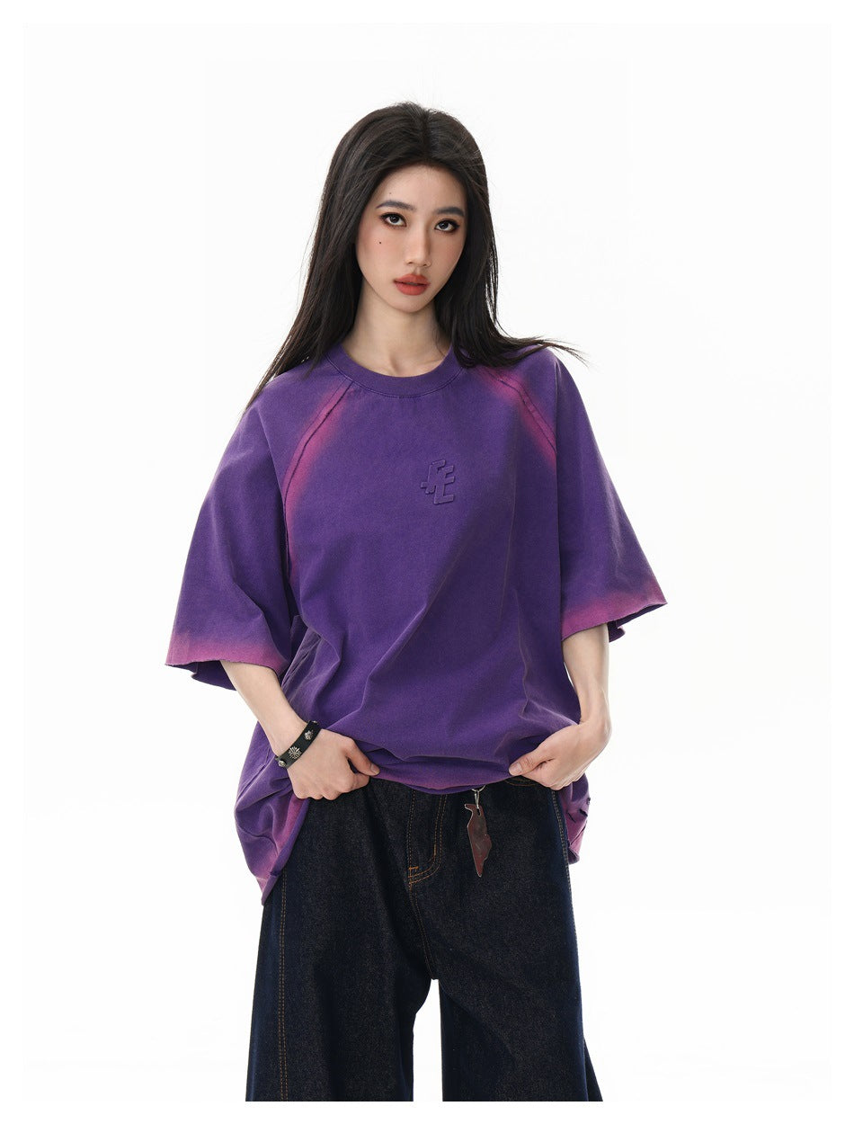 Model wearing a purple oversized T-shirt with pink faded accents and a small raised chest emblem.