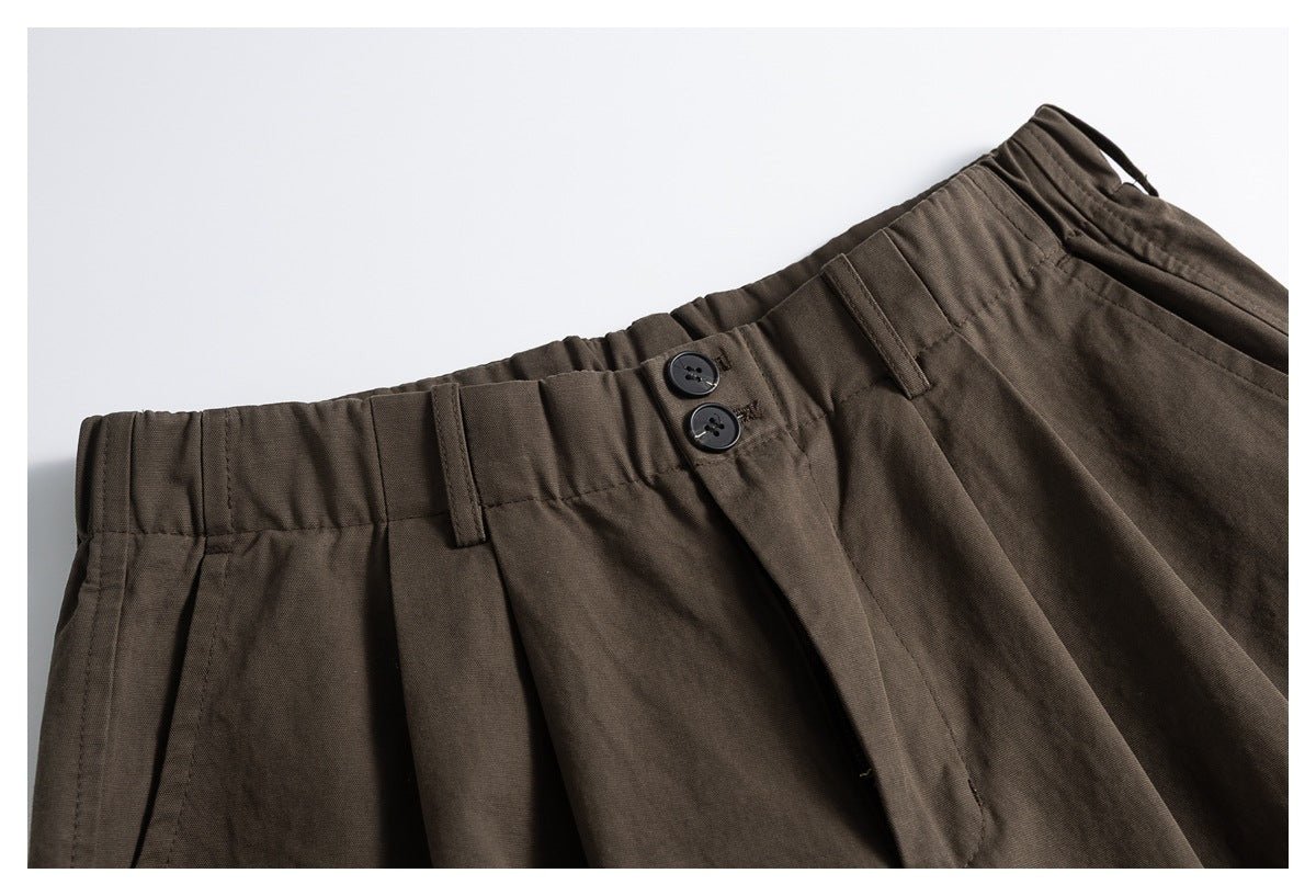 Close-up of brown trousers showing the gathered waistband, belt loops, and two-button front closure.