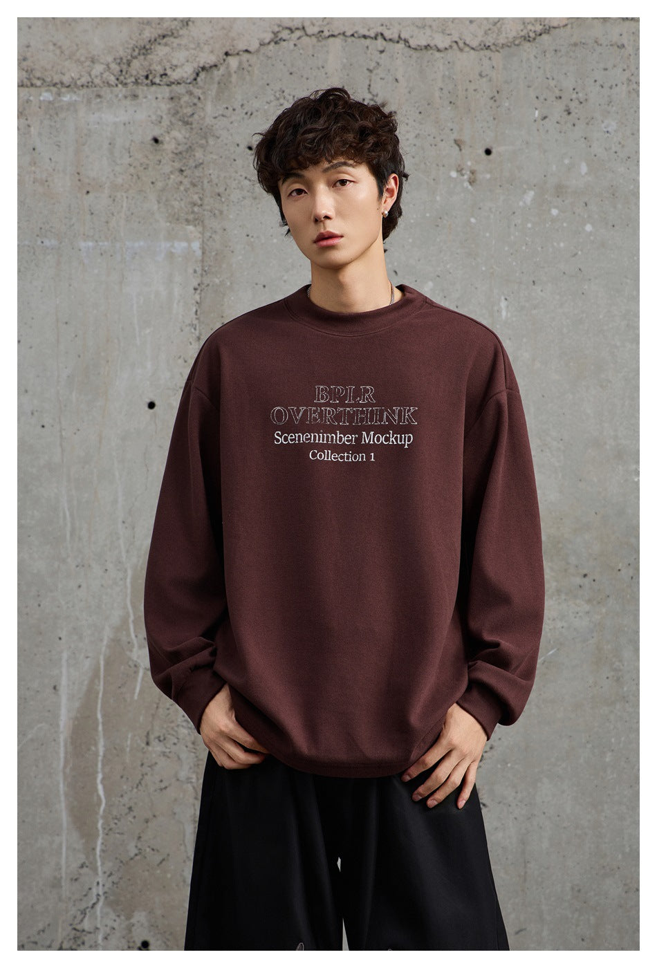 Model wearing a burgundy oversized crewneck sweatshirt with white chest text, shown from the front with black trousers.