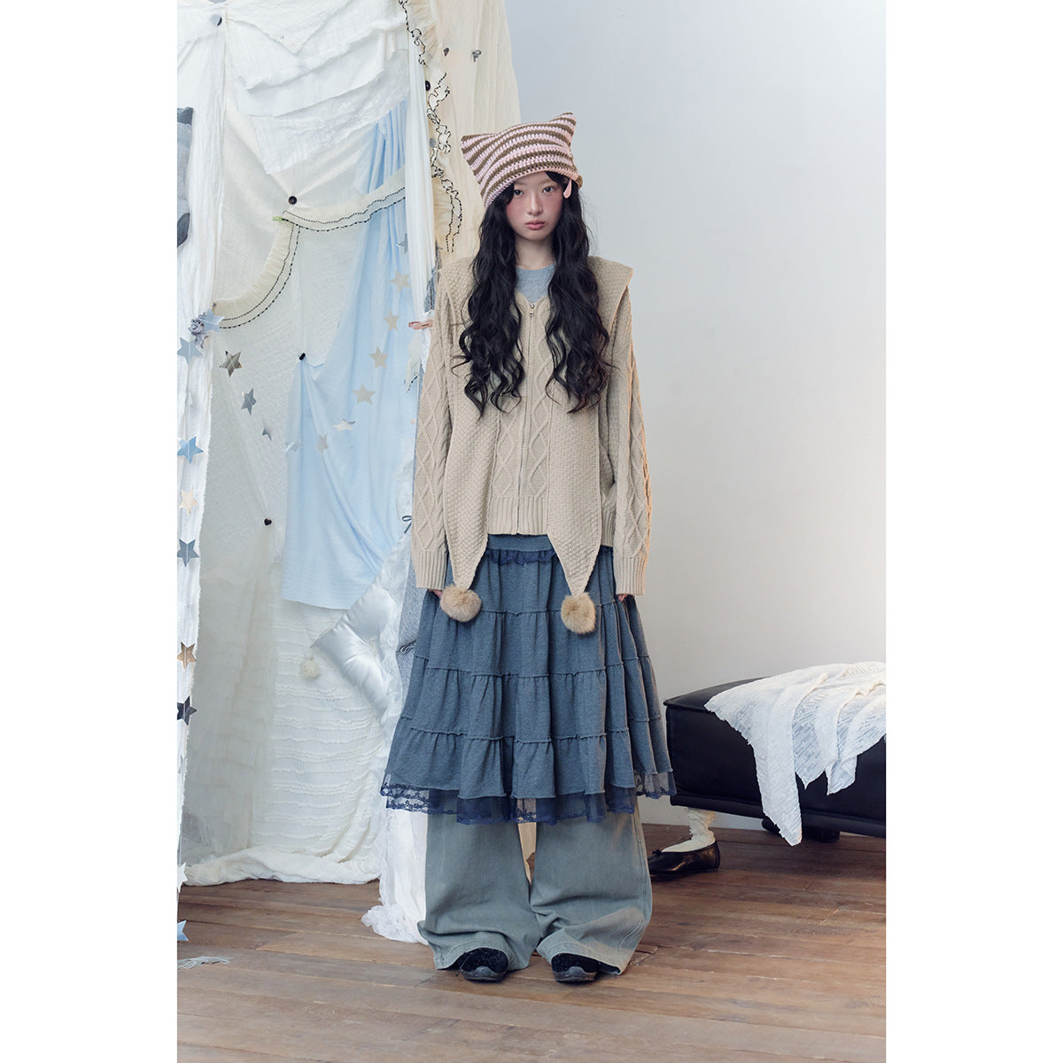 Full-length front view of a model wearing a beige cable-knit zip hoodie over a tiered blue skirt and wide-leg pants.