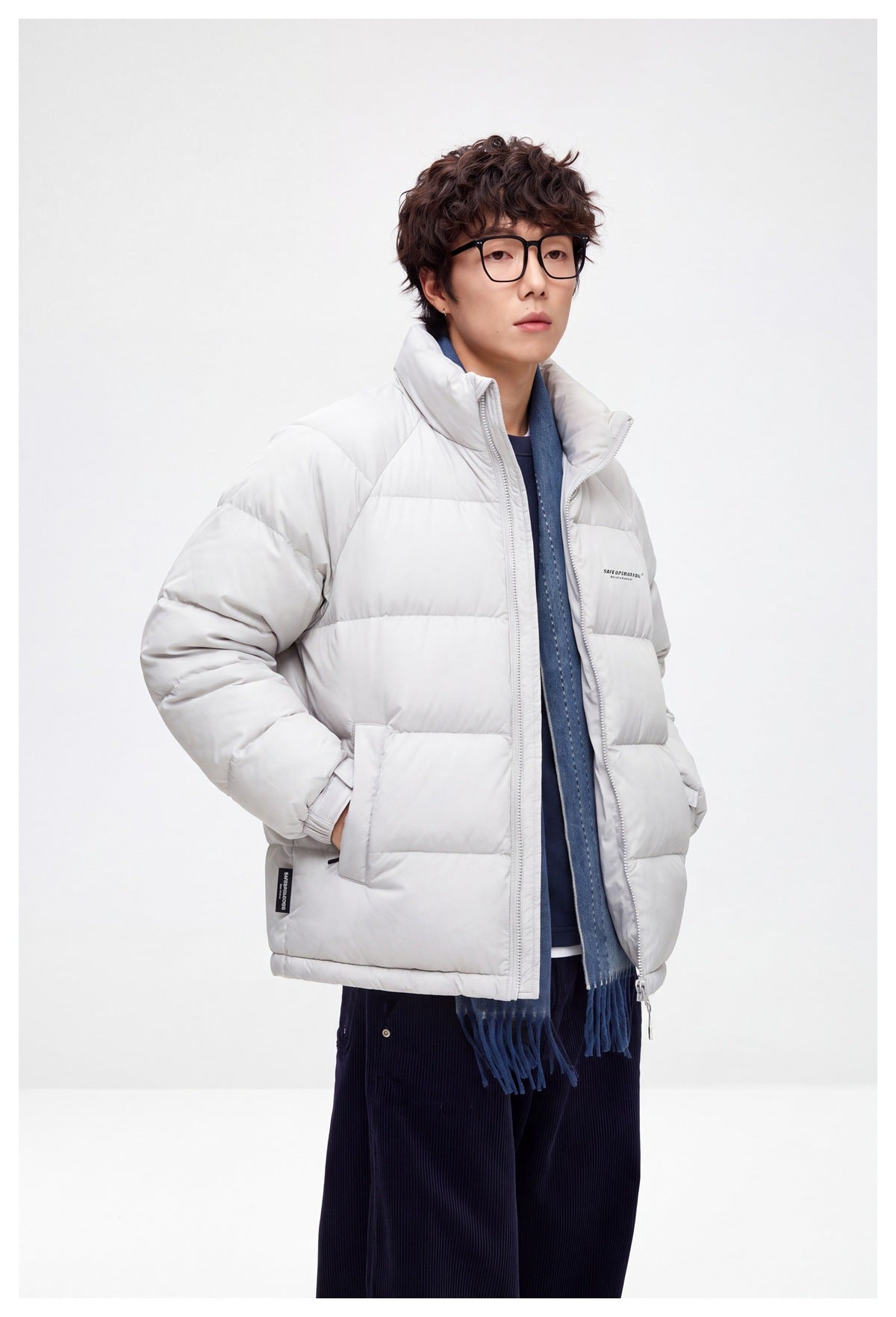 Model wearing a light gray quilted puffer jacket with a high collar, styled with a blue scarf and dark trousers.