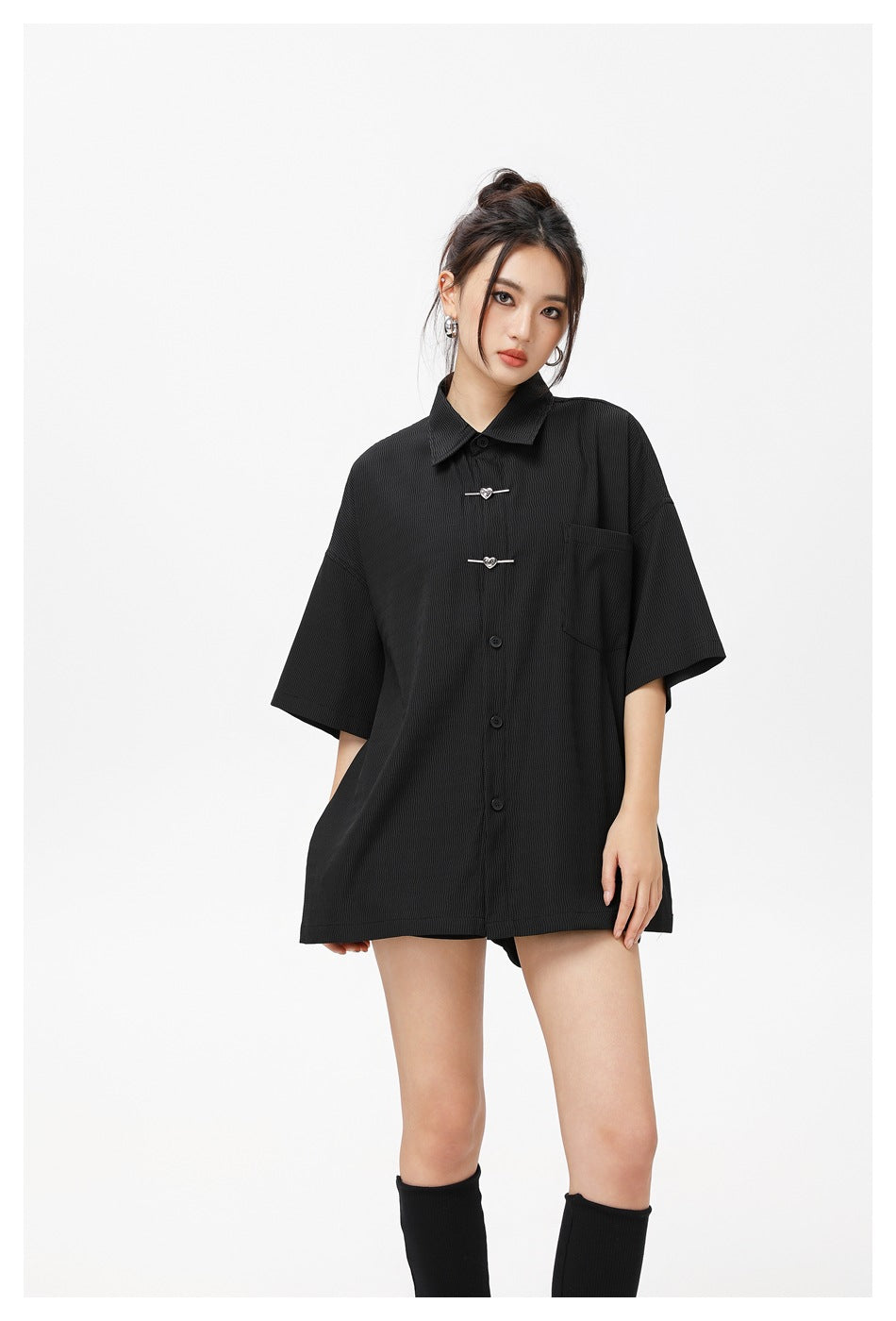 Model wearing a black oversized short-sleeve button-up shirt with a chest pocket and decorative metal toggles.