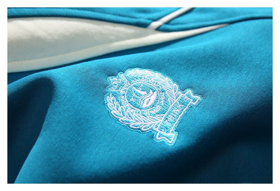 Close-up of a white embroidered crest on the teal sweatshirt, with a cream panel and white piping nearby.