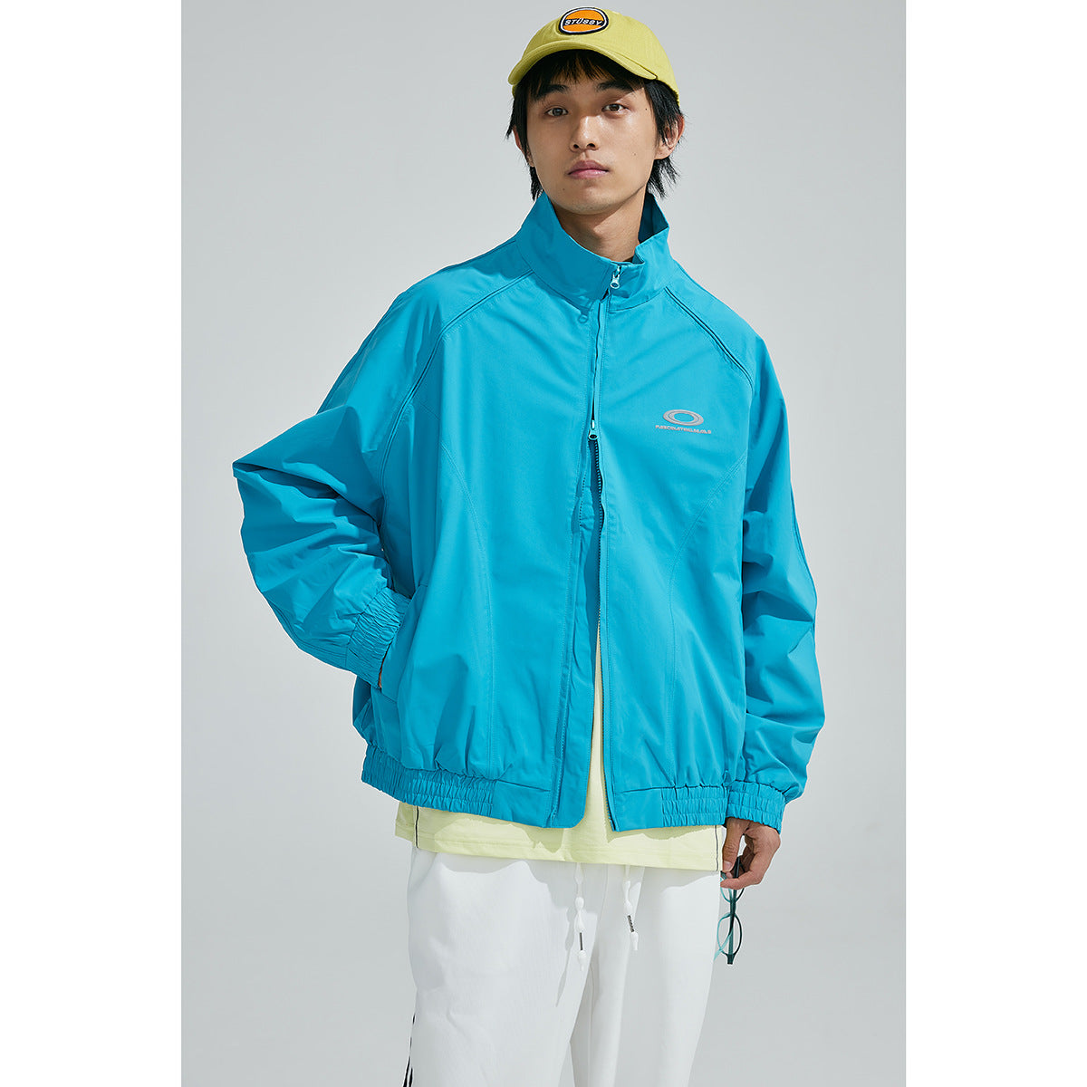 Model wearing a bright turquoise zip-up jacket with a high collar, elastic cuffs and hem, shown from the front.
