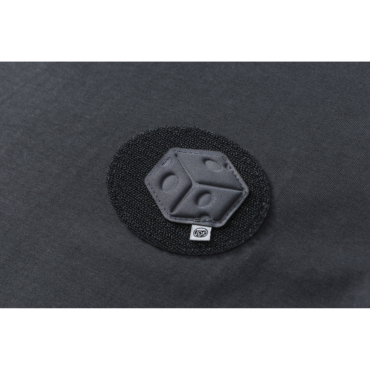 Close-up of a raised dark emblem patch on charcoal fabric, with a small symbol tab below it.