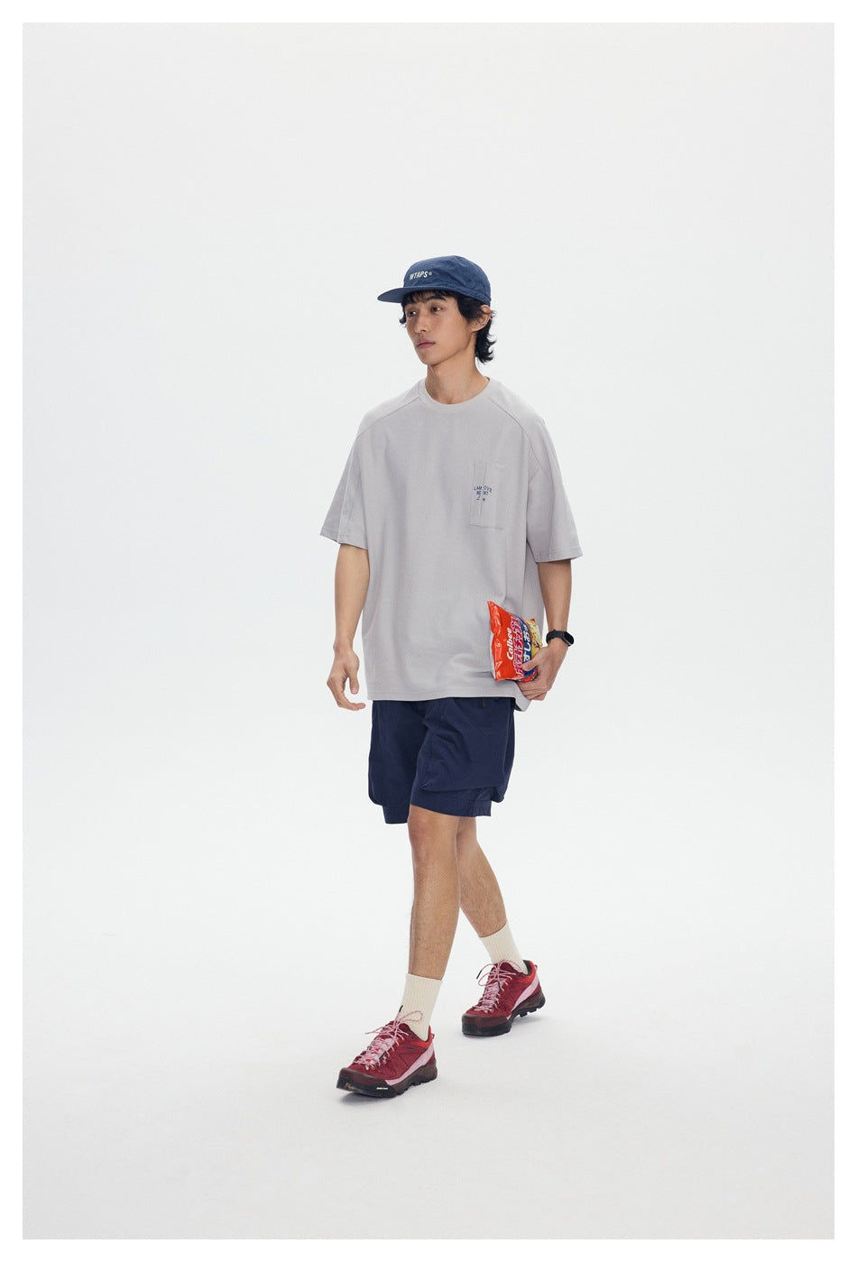 Model wearing a light gray pocket T-shirt, navy shorts, and cap while holding a snack bag.