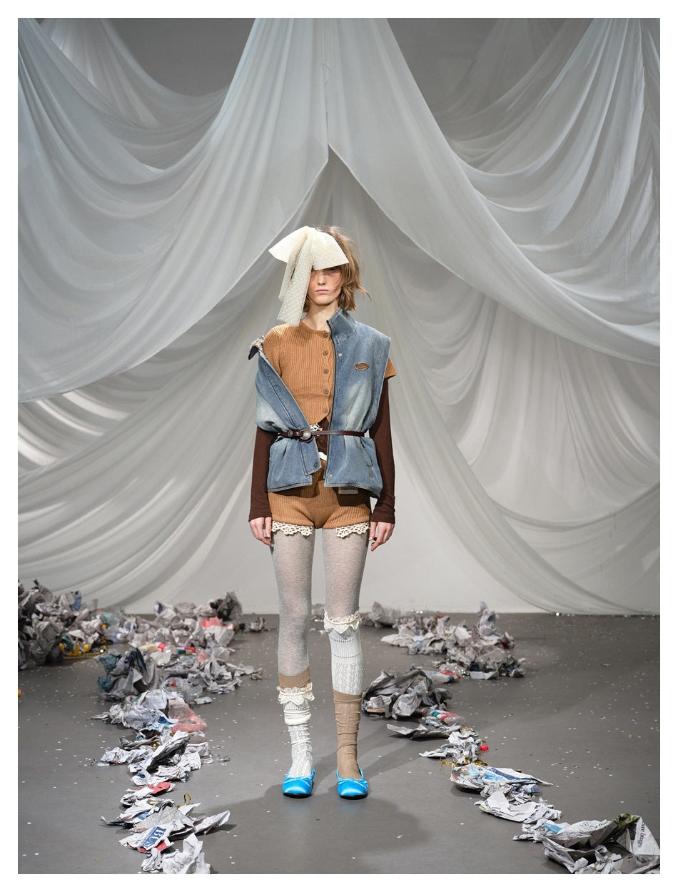 Person on a runway set wearing the denim vest open at the front over a ribbed top, belt, shorts and tights
