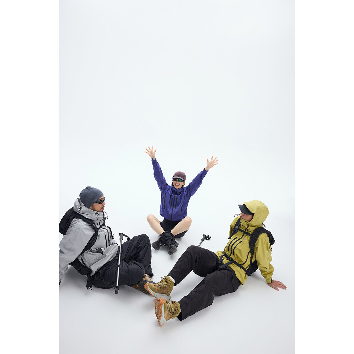 Three hikers wear gray, violet, and yellow hooded outdoor jackets while sitting together on a white background.