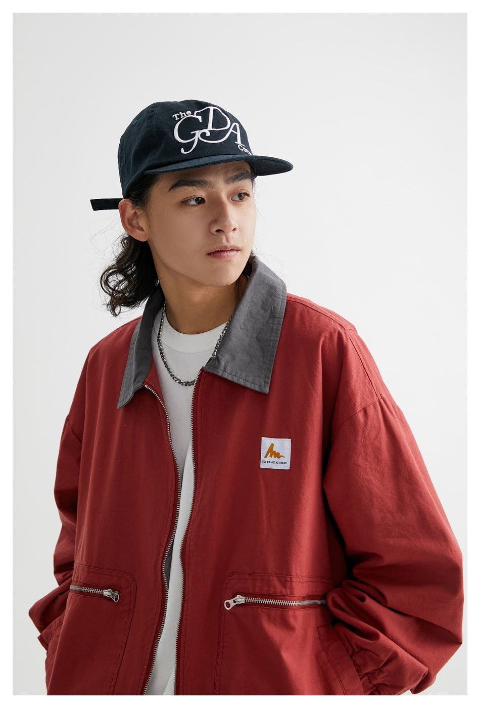 Close view of a model wearing a rust-red jacket with a gray collar, silver front zipper, and zippered pockets.