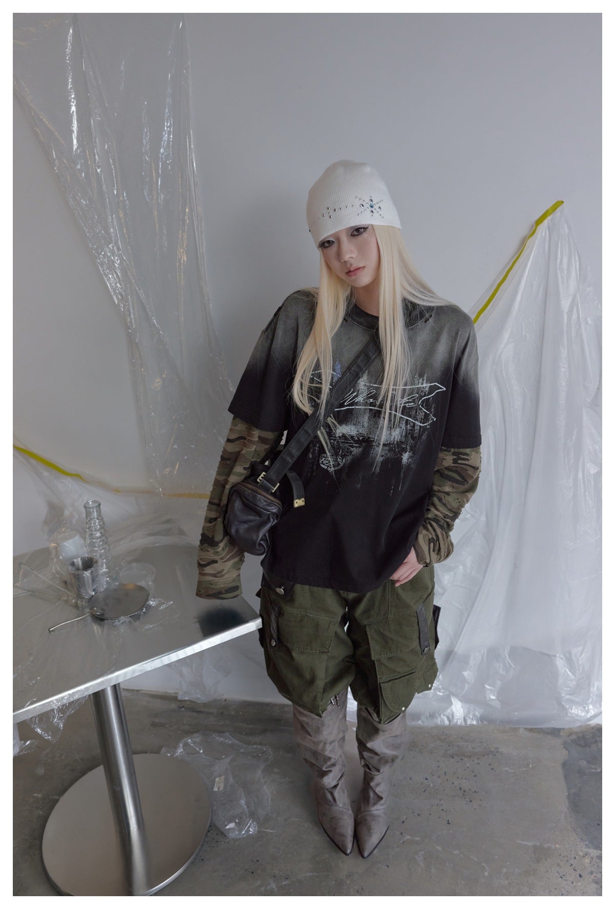 Model wearing an oversized black graphic tee over camouflage sleeves, olive cargo pants, a white beanie, and gray boots.