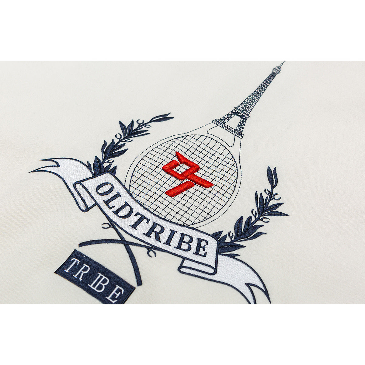 Close-up of an embroidered crest with an Eiffel Tower, tennis racket, laurel branches and text on ivory fabric.