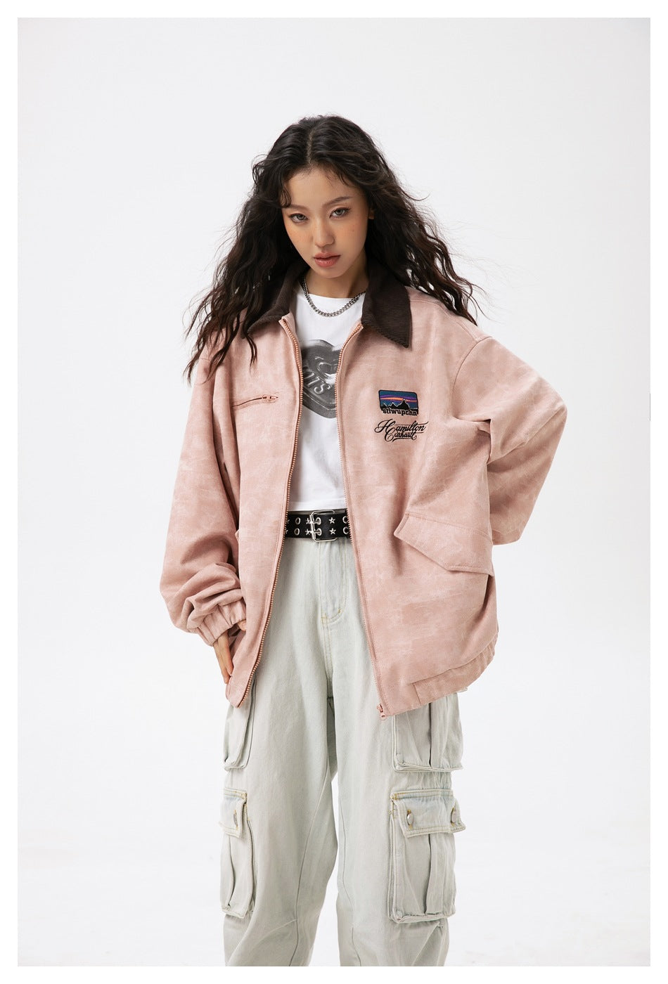 Model wearing a dusty pink zip jacket with dark brown collar and embroidered chest patch, paired with pale cargo pants.