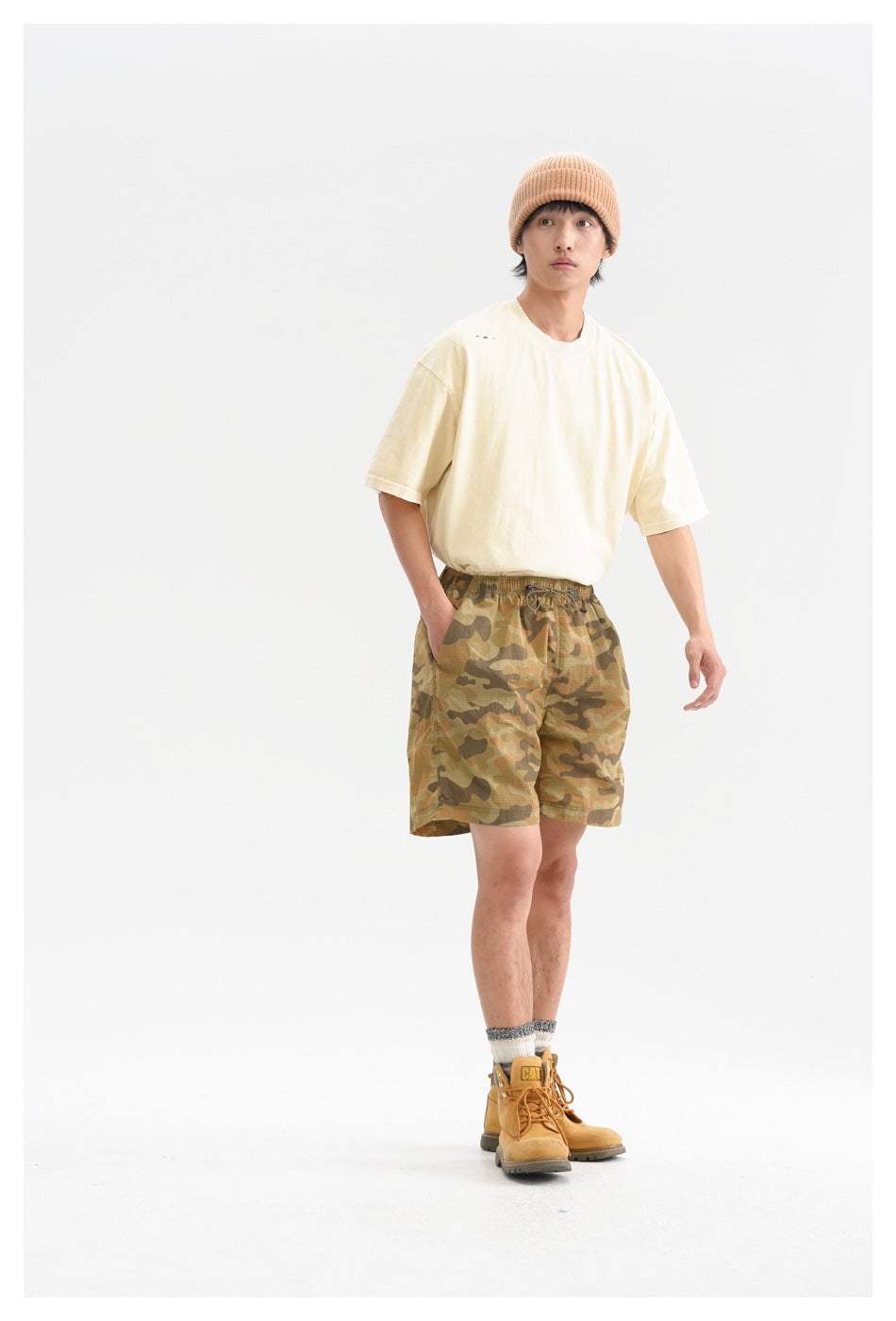 Man wearing a cream oversized T-shirt and tan camouflage drawstring shorts, standing against a white studio background.