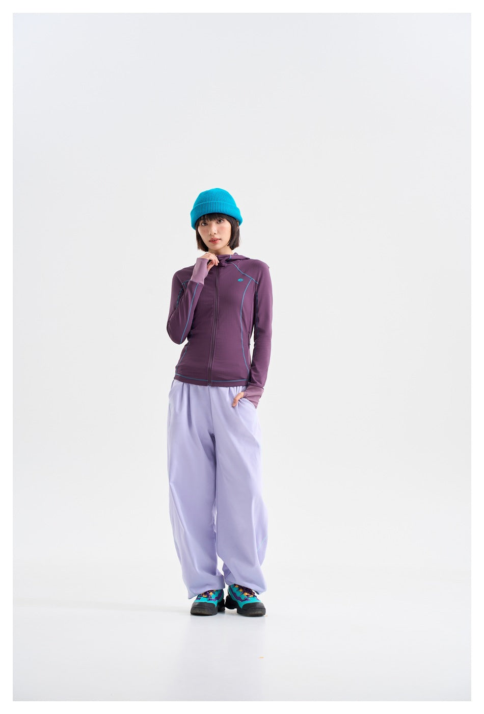 Model wearing a purple zip-up hooded jacket with turquoise piping, loose lavender pants, and a bright blue beanie.