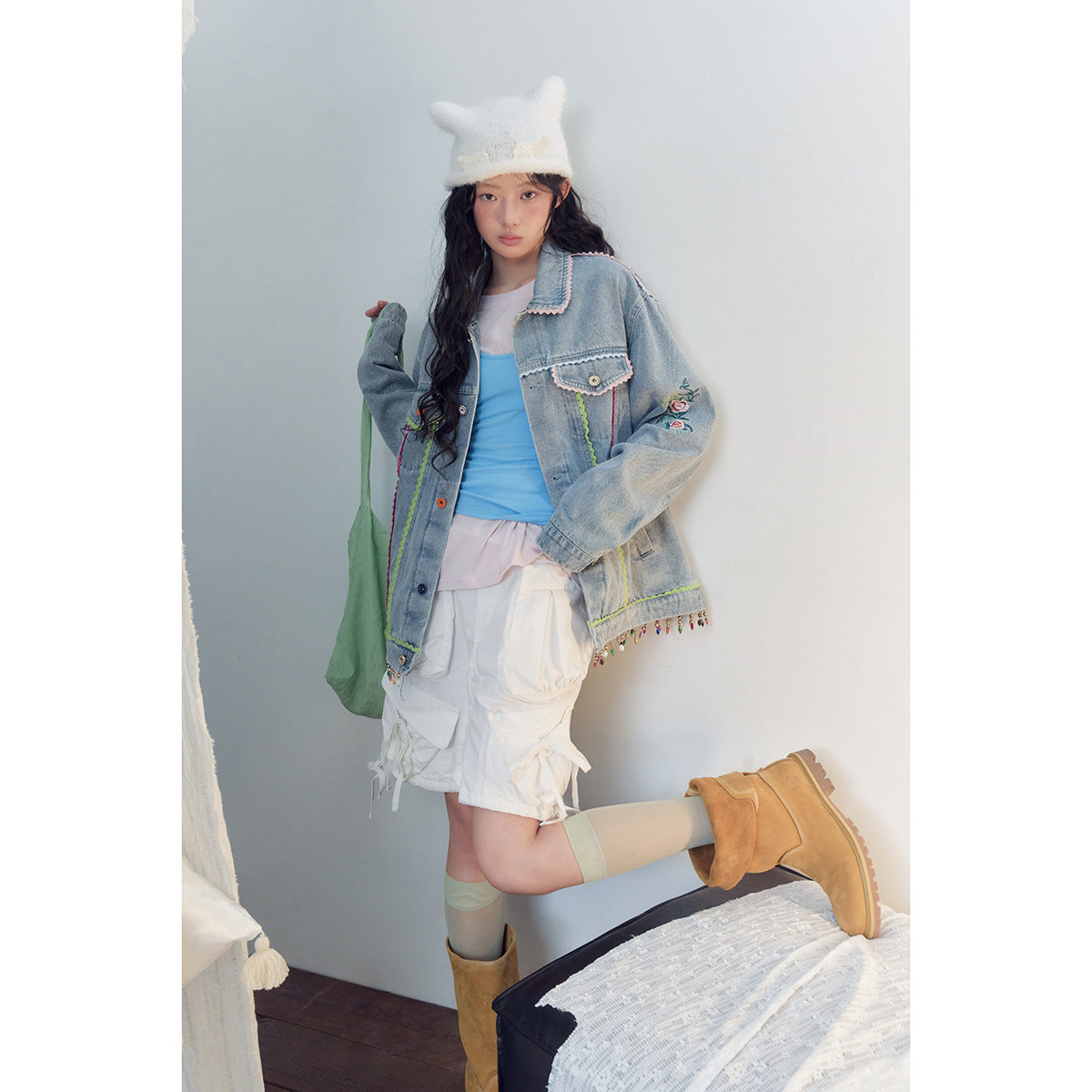 Model in a light-wash denim jacket with floral embroidery, bright trim, and dangling beads, paired with white shorts.